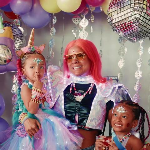 A parent with pink hair and two kids in tutus and face gems posing under balloons and a disco ball
