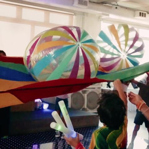 Kids with glow sticks under a rainbow parachute with giant beach balls